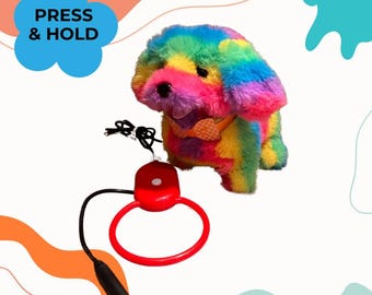 Peut inclure: Une peluche de chien aux couleurs de l'arc-en-ciel avec un petit nœud papillon orange. Le jouet est relié par un cordon noir à une manette circulaire rouge dotée d'un bouton blanc. Le texte PRESS & HOLD apparaît dans un nuage bleu.
