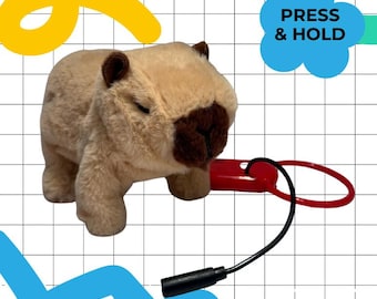 Peut inclure: Une peluche capybara beige et marron, avec une étiquette rouge reliée par un cordon noir. Le jouet présente des détails bruns et le texte "PRESS & HOLD" dans une bulle bleue.