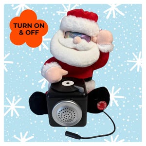May include: A plush Santa Claus figure wearing holographic sunglasses and a red suit with white trim. The figure is positioned behind a black square speaker base featuring a record player design on top and a silver speaker grille. Text reads TURN ON & OFF.
