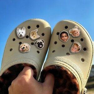 May include: Beige clogs decorated with charms depicting pets and children's faces. The clogs have a faux fur lining. The image shows the clogs held up against a blue sky.