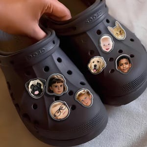 May include: Dark grey Crocs shoes decorated with face charms, including a cat, a baby, a golden retriever, and children. The charms are round and feature detailed images. The shoes are shown from a top-down perspective.