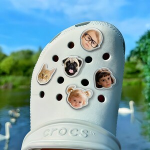 May include: A white Crocs shoe decorated with charms. The charms depict a cat, a pug, a child in glasses, and two other children. The shoe is positioned against a backdrop of a lake and a blue sky.