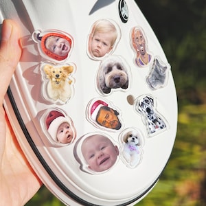 May include: A white shoe decorated with clear acrylic charms. The charms display images of babies, dogs, a cat, and a basketball player. The shoe is held by a hand, with a blurred green backdrop.