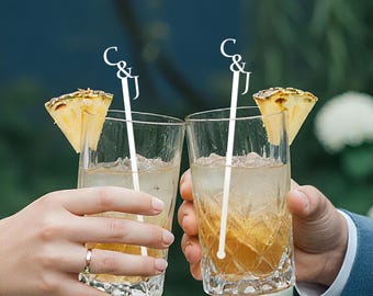May include: Two clear glasses filled with a light-colored beverage and ice, garnished with pineapple wedges. Each glass has a white stirrer with the letters "C & J". The glasses are held by two people, one wearing a ring.