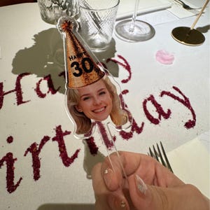 May include: A clear acrylic cake topper with a smiling woman's face and a gold glitter party hat with the number 30. The words "Happy Birthday" are written on the table in red.