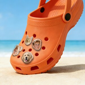May include: Orange clog shoe with decorative charms. The charms include a cat, a child's face, a dog, and another child's face. The shoe is on a sandy surface with a blue sky and ocean in the background.