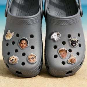 May include: Two grey clogs adorned with face charms. Charms feature a cat, a golden retriever, a young boy, a child with glasses, a pug, a dalmatian, and a baby. The clogs are set on a sandy surface with a blue sky backdrop.