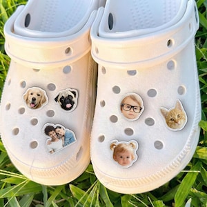 May include: White clogs decorated with custom charms. The charms display images of a golden retriever, a pug, a couple, a child wearing glasses, a cat, and a baby with pigtails. The clogs are on green grass.