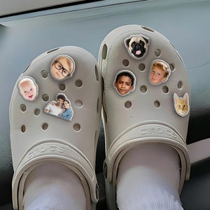 May include: A pair of light gray Crocs adorned with various decorative charms featuring faces of people and pets. The charms include a baby, a child wearing glasses, a couple, a pug, a young boy, and a cat. The Crocs are worn with white socks.