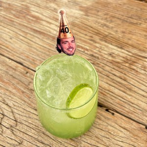 May include: A green cocktail in a clear glass with ice and a lime wedge. A small party hat with the text "Happy 40th" and a man's face is attached to the top of the glass. The drink sits on a wooden surface.