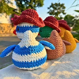 Rear view of three crocheted duck dolls standing in a row on a light-colored cloth surface outdoors. The ducks are wearing maroon cowboy hats. The nearest duck has blue and white stripes, the middle one has a green head and brown body, and the farthest one is orange. The background includes a quiet street, trees, and greenery in soft evening sunlight.