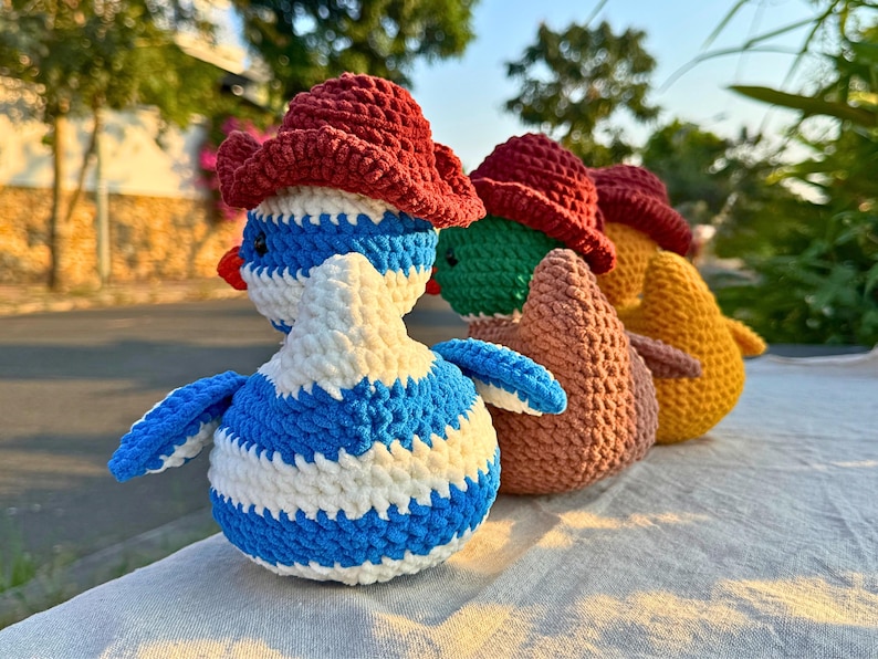 Rear view of three crocheted duck dolls standing in a row on a light-colored cloth surface outdoors. The ducks are wearing maroon cowboy hats. The nearest duck has blue and white stripes, the middle one has a green head and brown body, and the farthest one is orange. The background includes a quiet street, trees, and greenery in soft evening sunlight.