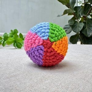 K&ouml;nnte beinhalten: Ein farbenfroher, geh&auml;kelter Ball mit Segmenten in Blau, Rosa, Lila, Rot, Orange und Gr&uuml;n. Der handgefertigte Ball liegt auf einer neutralfarbenen Oberfl&auml;che, mit gr&uuml;nem Laub im Hintergrund.