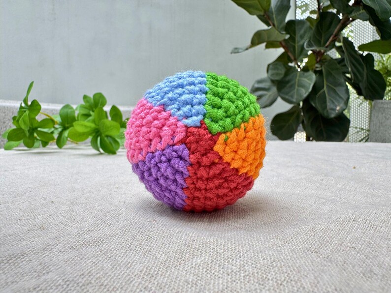 K&ouml;nnte beinhalten: Ein farbenfroher, geh&auml;kelter Ball mit Segmenten in Blau, Rosa, Lila, Rot, Orange und Gr&uuml;n. Der handgefertigte Ball liegt auf einer neutralfarbenen Oberfl&auml;che, mit gr&uuml;nem Laub im Hintergrund.