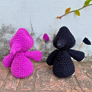 May include: Two crocheted plush toys, one in pink and one in black, holding sticks with small flags on the ends. The toys are standing on a brick surface.