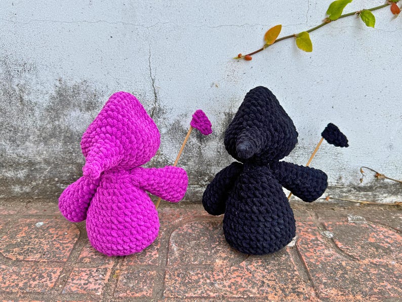 May include: Two crocheted plush toys, one in pink and one in black, holding sticks with small flags on the ends. The toys are standing on a brick surface.