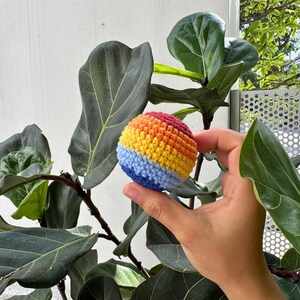 K&ouml;nnte beinhalten: Ein kleiner, runder, geh&auml;kelter Ball mit Regenbogenfarben. Der Ball hat B&auml;nder in Blau, Gelb, Orange und Rot. Der Ball wird vor einer gro&szlig;bl&auml;ttrigen Pflanze gehalten.