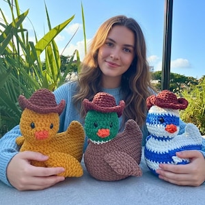 May include: Three crocheted duck toys, each wearing a brown cowboy hat. One is yellow, one is green and brown, and the third is white and blue. The ducks have orange beaks and are sitting on a light blue surface.