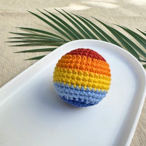 K&ouml;nnte beinhalten: Ein geh&auml;kelter Ball in Regenbogenfarben: Rot, Orange, Gelb und Blau. Der Ball liegt auf einem wei&szlig;en, oval geformten Tablett. Der Hintergrund umfasst eine beige Oberfl&auml;che und ein gr&uuml;nes Palmenblatt.