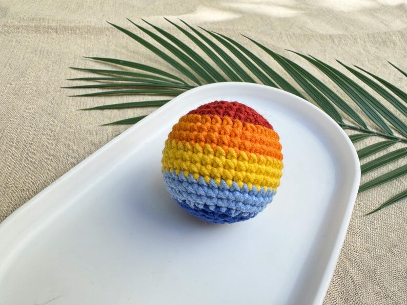 K&ouml;nnte beinhalten: Ein geh&auml;kelter Ball in Regenbogenfarben: Rot, Orange, Gelb und Blau. Der Ball liegt auf einem wei&szlig;en, oval geformten Tablett. Der Hintergrund umfasst eine beige Oberfl&auml;che und ein gr&uuml;nes Palmenblatt.