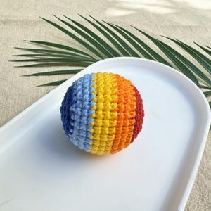 K&ouml;nnte beinhalten: Ein mehrfarbiger, geh&auml;kelter Ball mit blauen, gelben, orangefarbenen und roten Segmenten. Der Ball liegt auf einem wei&szlig;en, ovalen Teller. Der Hintergrund ist neutral. Die Farben des Balls sind in vertikalen Streifen angeordnet.