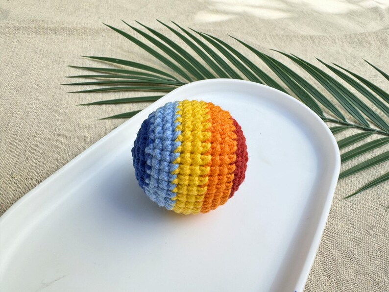 K&ouml;nnte beinhalten: Ein mehrfarbiger, geh&auml;kelter Ball mit blauen, gelben, orangefarbenen und roten Segmenten. Der Ball liegt auf einem wei&szlig;en, ovalen Teller. Der Hintergrund ist neutral. Die Farben des Balls sind in vertikalen Streifen angeordnet.