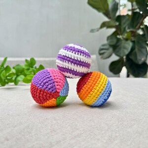 K&ouml;nnte beinhalten: Drei bunte H&auml;kelb&auml;lle. Ein Ball ist lila und wei&szlig; gestreift. Ein anderer ist ein Patchwork aus Rot, Orange, Gr&uuml;n, Blau, Rosa und Lila. Der dritte Ball ist mit einem Regenbogen aus Farben gestreift. Die B&auml;lle liegen auf einer hellen Oberfl&auml;che.