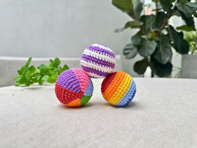 K&ouml;nnte beinhalten: Drei bunte H&auml;kelb&auml;lle. Ein Ball ist lila und wei&szlig; gestreift. Ein anderer ist ein Patchwork aus Rot, Orange, Gr&uuml;n, Blau, Rosa und Lila. Der dritte Ball ist mit einem Regenbogen aus Farben gestreift. Die B&auml;lle liegen auf einer hellen Oberfl&auml;che.