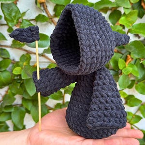 May include: Black crocheted amigurumi figure with a pointed hood, holding a small black flag on a wooden stick. The figure is standing on a person's hand.