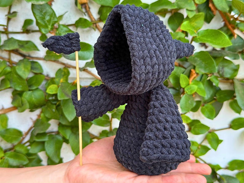May include: Black crocheted amigurumi figure with a pointed hood, holding a small black flag on a wooden stick. The figure is standing on a person's hand.