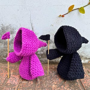 Könnte beinhalten: Zwei gehäkelte Figuren mit Kapuze, eine in Pink und eine in Schwarz, die jeweils eine kleine Flagge halten. Die Figuren stehen auf einer Ziegelstein-Oberfläche.