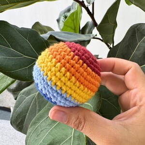 K&ouml;nnte beinhalten: Ein kleiner, runder, geh&auml;kelter Ball in den Farben des Regenbogens: Blau, Gelb, Orange und Rot. Der Ball wird vor einem Hintergrund aus gro&szlig;en, gr&uuml;nen Bl&auml;ttern gehalten.