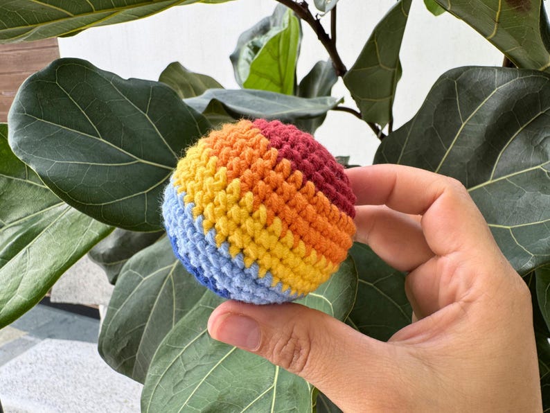 K&ouml;nnte beinhalten: Ein kleiner, runder, geh&auml;kelter Ball in den Farben des Regenbogens: Blau, Gelb, Orange und Rot. Der Ball wird vor einem Hintergrund aus gro&szlig;en, gr&uuml;nen Bl&auml;ttern gehalten.