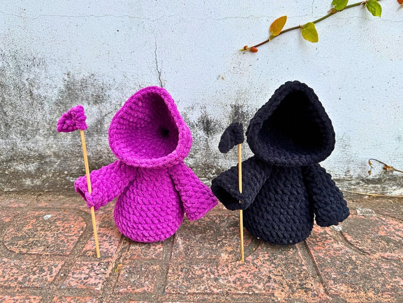 May include: Two crocheted hooded figures, one in pink and one in black, holding wooden sticks with small flags at the top.