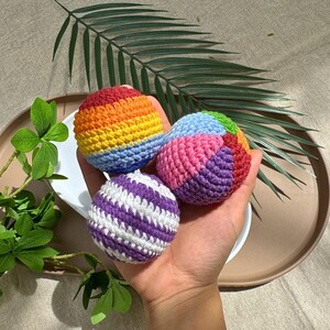 K&ouml;nnte beinhalten: Drei farbenfrohe geh&auml;kelte B&auml;lle. Einer ist lila und wei&szlig; gestreift, ein anderer hat Regenbogenfarben und der dritte ist rosa, blau, gr&uuml;n und orange. Die B&auml;lle sind in einer Hand, auf einem wei&szlig;en Teller und einem braunen Tablett, mit gr&uuml;nen Bl&auml;ttern im Hintergrund.