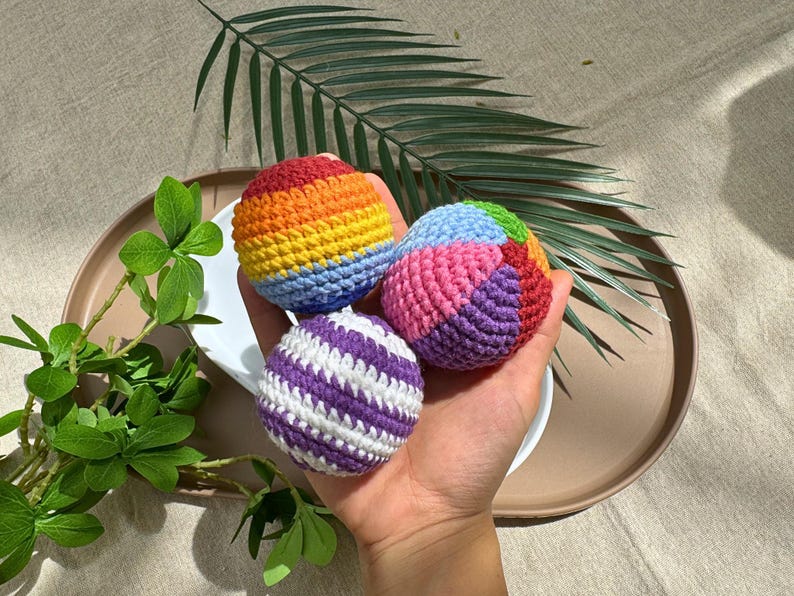 K&ouml;nnte beinhalten: Drei farbenfrohe geh&auml;kelte B&auml;lle. Einer ist lila und wei&szlig; gestreift, ein anderer hat Regenbogenfarben und der dritte ist rosa, blau, gr&uuml;n und orange. Die B&auml;lle sind in einer Hand, auf einem wei&szlig;en Teller und einem braunen Tablett, mit gr&uuml;nen Bl&auml;ttern im Hintergrund.