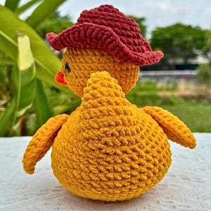 Pu&ograve; includere: Un pulcino giallo fatto a mano all'uncinetto con un cappello da cowboy rosso. Il pulcino ha accenti arancioni per il becco e le zampe. Il peluche &egrave; appoggiato su una superficie bianca con fogliame verde sullo sfondo.