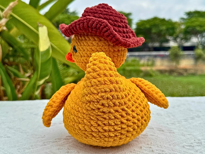 Pu&ograve; includere: Un pulcino giallo fatto a mano all'uncinetto con un cappello da cowboy rosso. Il pulcino ha accenti arancioni per il becco e le zampe. Il peluche &egrave; appoggiato su una superficie bianca con fogliame verde sullo sfondo.