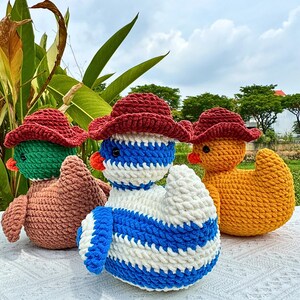 Pu&ograve; includere: Tre anatre all'uncinetto fatte a mano con cappelli da cowboy rossi. Una &egrave; verde e marrone, un'altra blu e bianca e la terza gialla. Sono su una superficie bianca con vegetazione sullo sfondo.