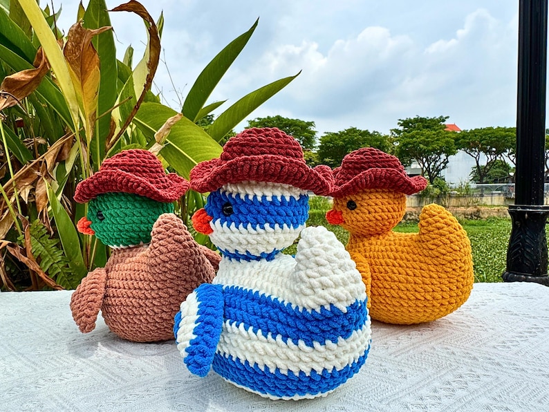 Pu&ograve; includere: Tre anatre all'uncinetto fatte a mano con cappelli da cowboy rossi. Una &egrave; verde e marrone, un'altra blu e bianca e la terza gialla. Sono su una superficie bianca con vegetazione sullo sfondo.