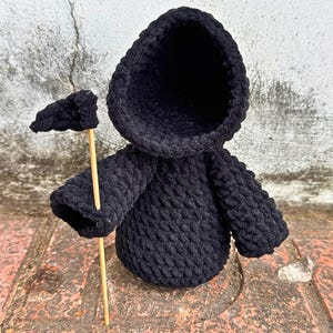 May include: Black crocheted hooded figure holding a small black flag. The figure is standing on a brick surface.