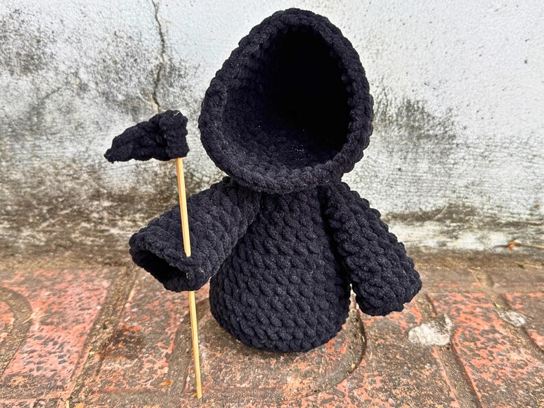 May include: Black crocheted hooded figure holding a small black flag. The figure is standing on a brick surface.
