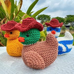 Pu&ograve; includere: Tre figure di anatra all'uncinetto con cappelli da cowboy rossi. Uno &egrave; giallo, uno &egrave; verde e uno &egrave; blu e bianco. Ogni anatra ha un becco arancione e occhi neri. Le anatre sono fatte a mano e sono su una superficie bianca.
