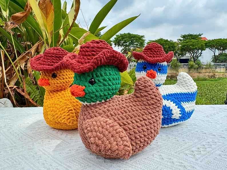 Pu&ograve; includere: Tre figure di anatra all'uncinetto con cappelli da cowboy rossi. Uno &egrave; giallo, uno &egrave; verde e uno &egrave; blu e bianco. Ogni anatra ha un becco arancione e occhi neri. Le anatre sono fatte a mano e sono su una superficie bianca.