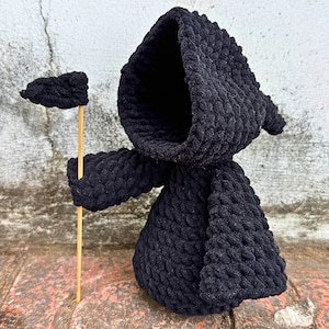 May include: Black crocheted hooded figure holding a small black flag on a wooden stick. The figure is standing on a brick surface.