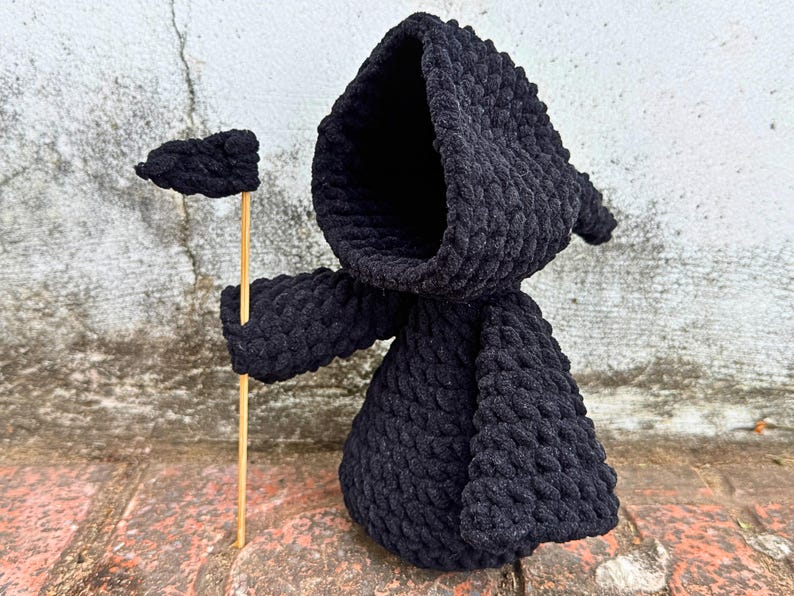 May include: Black crocheted hooded figure holding a small black flag on a wooden stick. The figure is standing on a brick surface.