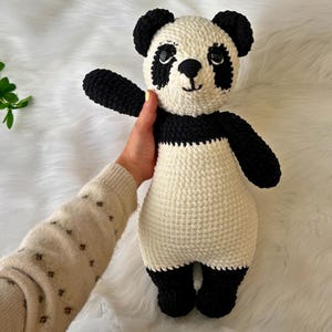 Op de afbeelding: Een handgemaakte gehaakte panda beer in zwart en wit. De panda heeft zwarte oren, ogen, armen en benen, met een witte body. De panda houdt een arm omhoog. De achtergrond is een wit, pluizig oppervlak.