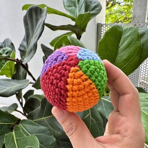 K&ouml;nnte beinhalten: Ein mehrfarbiger, geh&auml;kelter Ball mit Segmenten in Rot, Orange, Gr&uuml;n, Blau und Lila. Der Ball wird in einer Hand gehalten, mit einem Hintergrund aus gro&szlig;en gr&uuml;nen Bl&auml;ttern.