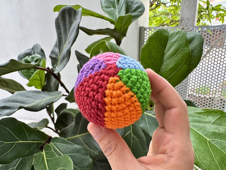 K&ouml;nnte beinhalten: Ein mehrfarbiger, geh&auml;kelter Ball mit Segmenten in Rot, Orange, Gr&uuml;n, Blau und Lila. Der Ball wird in einer Hand gehalten, mit einem Hintergrund aus gro&szlig;en gr&uuml;nen Bl&auml;ttern.