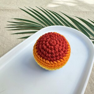 K&ouml;nnte beinhalten: Ein handgeh&auml;kelter Ball in Orange- und Gelbt&ouml;nen, der auf einem wei&szlig;en, ovalen Teller liegt. Die Textur des Balls ist sichtbar, mit einem Farbverlauf. Ein gr&uuml;nes Palmenblatt befindet sich im Hintergrund.